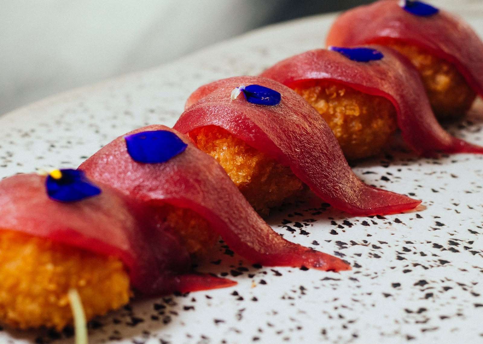 Croquetas doradas de langostino con láminas de sashimi de atún rojo y pétalos azules sobre un plato blanco