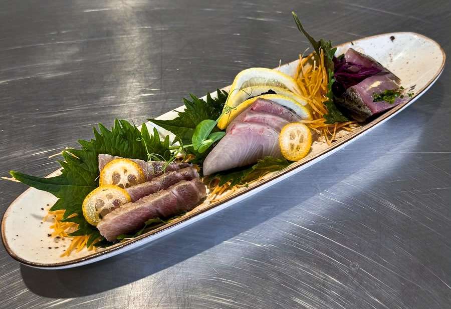 Fuente alargada con sashimi de o-toro, akami y kinmedai, cítricos y hojas sobre una mesa de acero